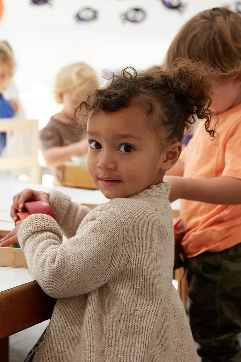 A preschooler looks toward the camera while classmates work at wooden tables