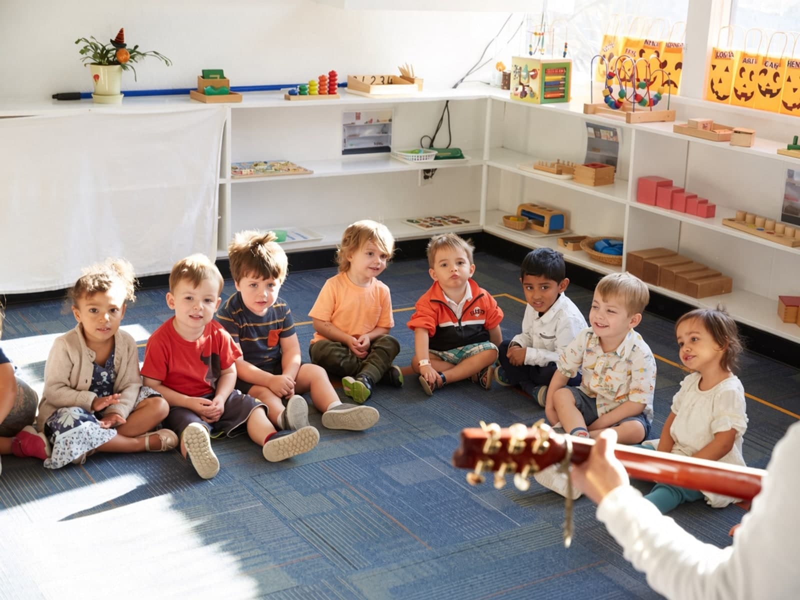 Preschoolers smile gently during classroom circle time while a teacher plays guitar
