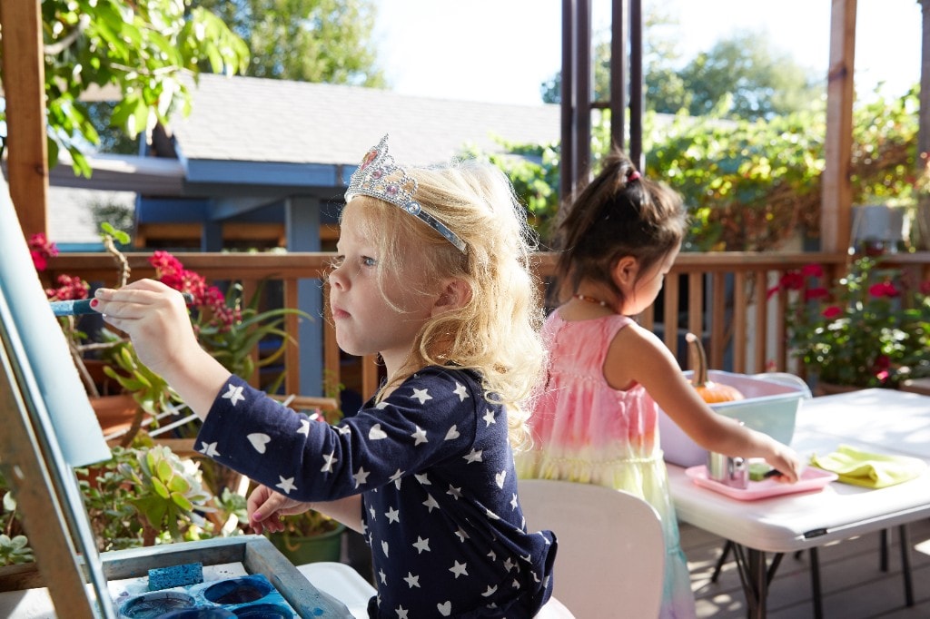 A child in a tiara paints at an easel on the sunny deck while a friend works at a table